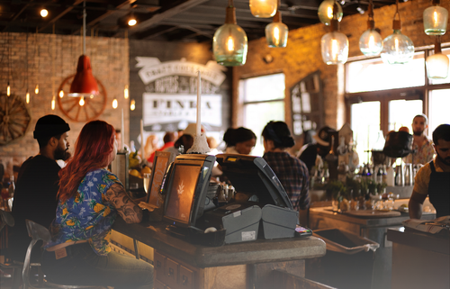 A warm, bustling restaurant interior with people seated at a bar and working behind the counter, surrounded by hanging pendant lights, rustic brick walls, and modern point‑of‑sale systems.